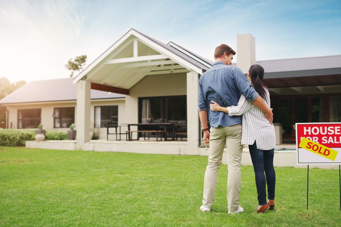 Two people standing in front of their new home, with a sold sign in the front.
